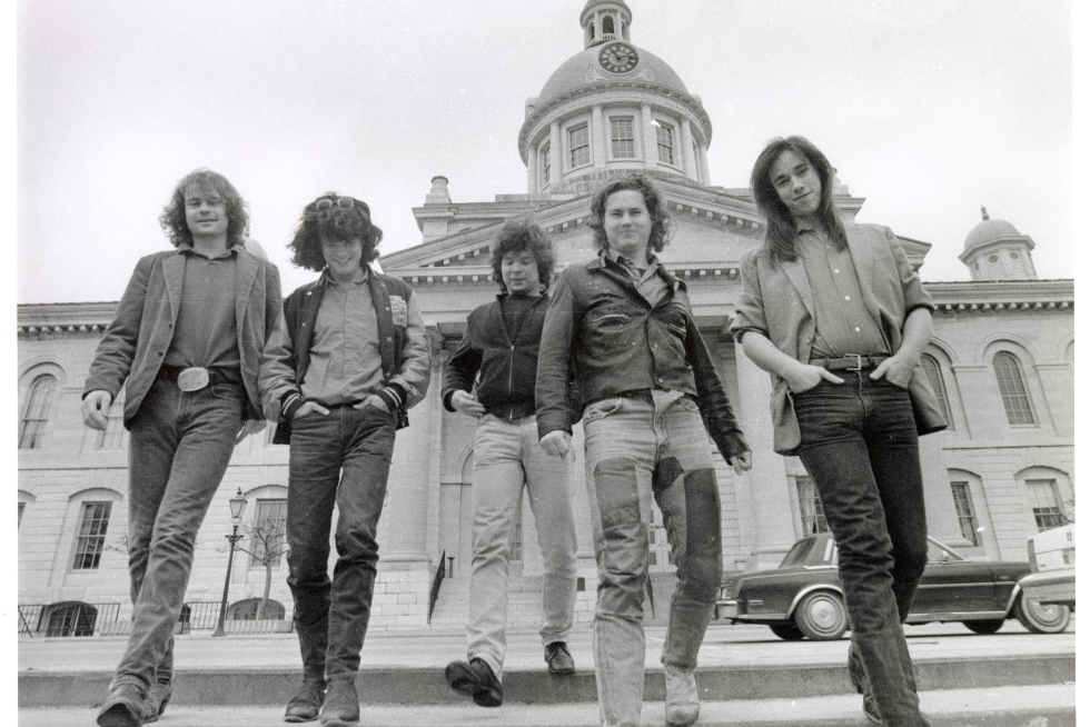 Members of the Tragically Hip stand smiling. Kingston City Hall is visible in the background.