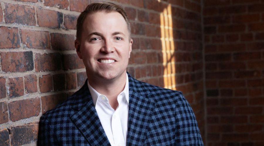 Graham Coe, MBA’18, dressed in a plaid jacket and white shirt, stands confidently before a brick wall backdrop.