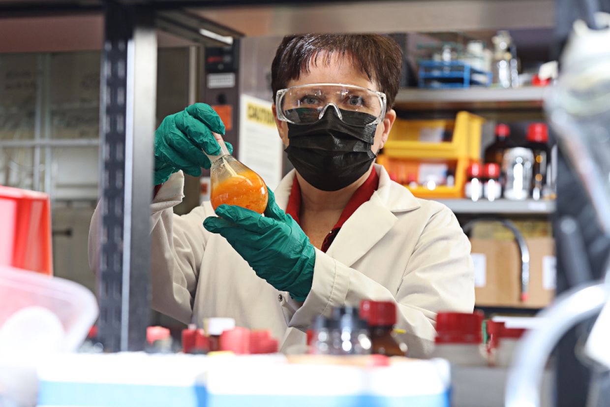 Cathleen Crudden in a lab coat, goggles, and mask holds a science beaker. 