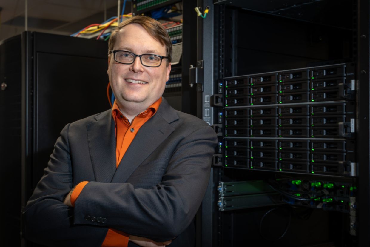 Dr. Ryan Grant, wearing glasses, stands in front of a server rack, showcasing his expertise in technology and research.