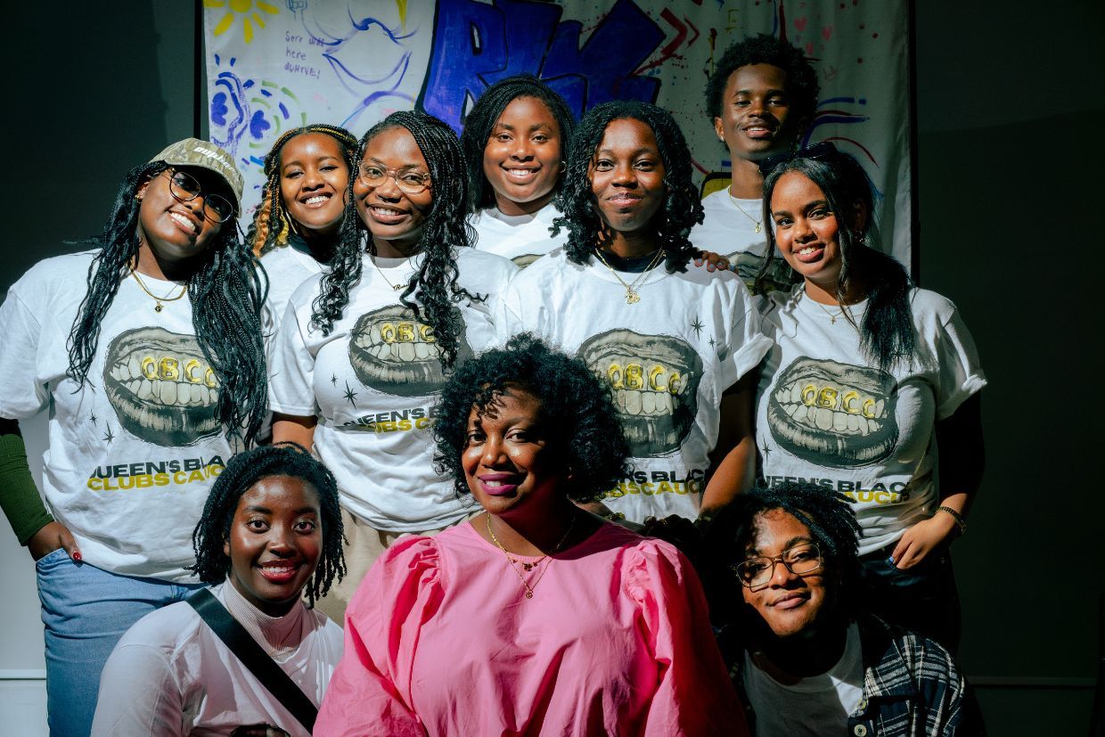 A group of people posing for a photo indoors, smiling and standing together against a wall. They are dressed casually in jeans and exhibit a sense of friendship and happiness.