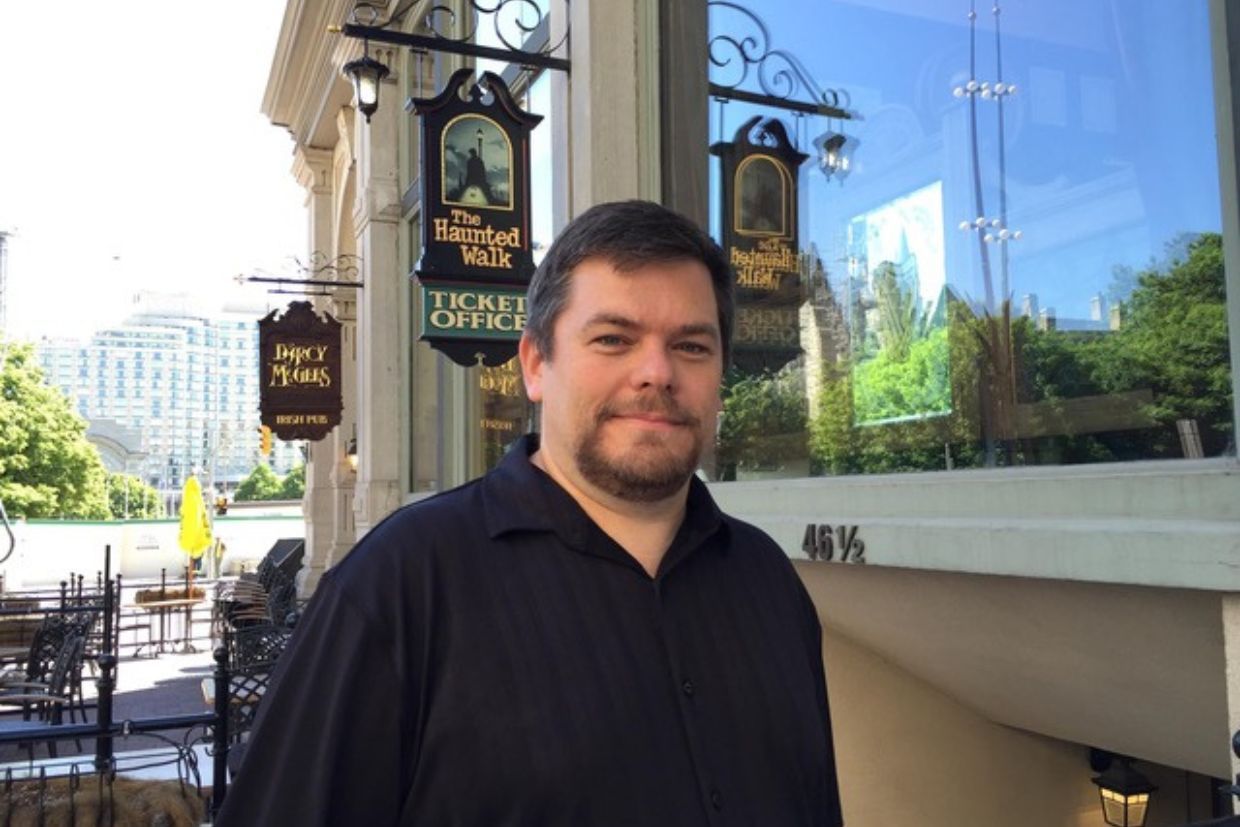 Glen Shackleton, Artsci’96, dressed in black, stands before a building displaying a sign for The Haunted Walk in Kingston.
