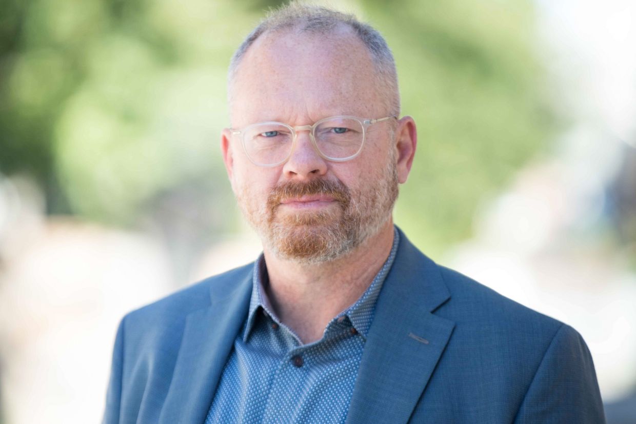 Headshot of Christopher Donaldson. He wears a blue suit and there is green foliage behind him. 