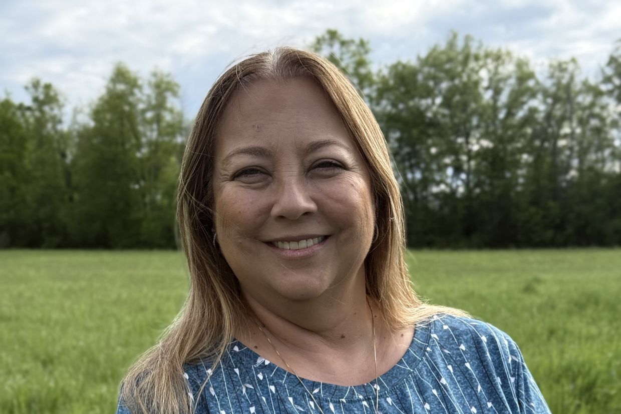 Lisa Maracle smiling, wearing a blue shirt, with a green-grass field behind her and trees beyond that.