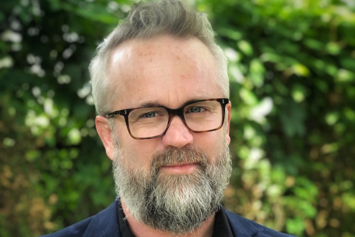 Headshot of Ron Shore wearing a blue jacket and dark glasses, with green vegetation behind him.