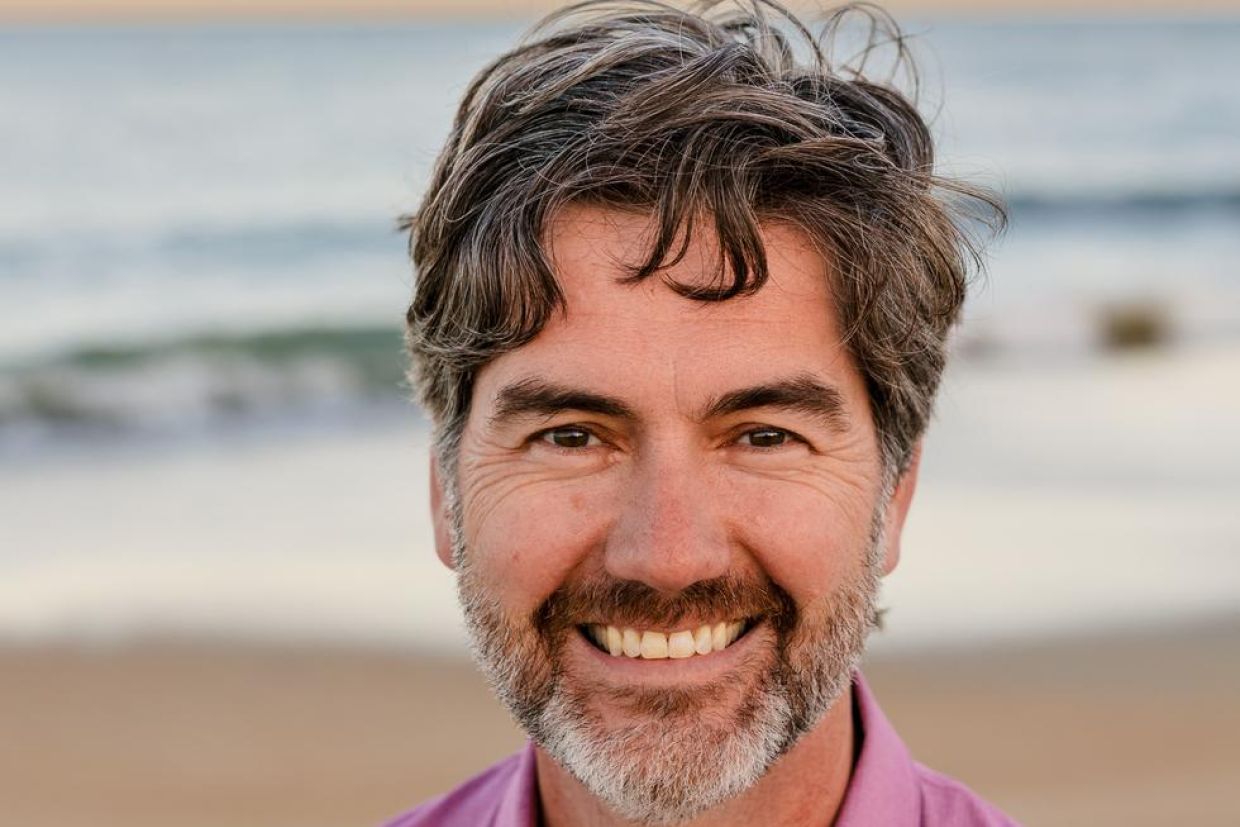 Close up of Ryan Mulligan's smiling face, with a beach and ocean behind him.