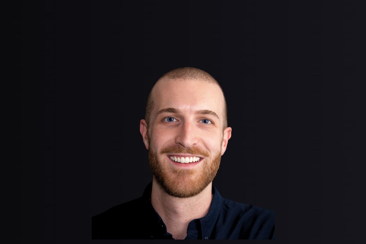 A smiling Sean McCabe wearing a dark-collared shirt. The background is black. 