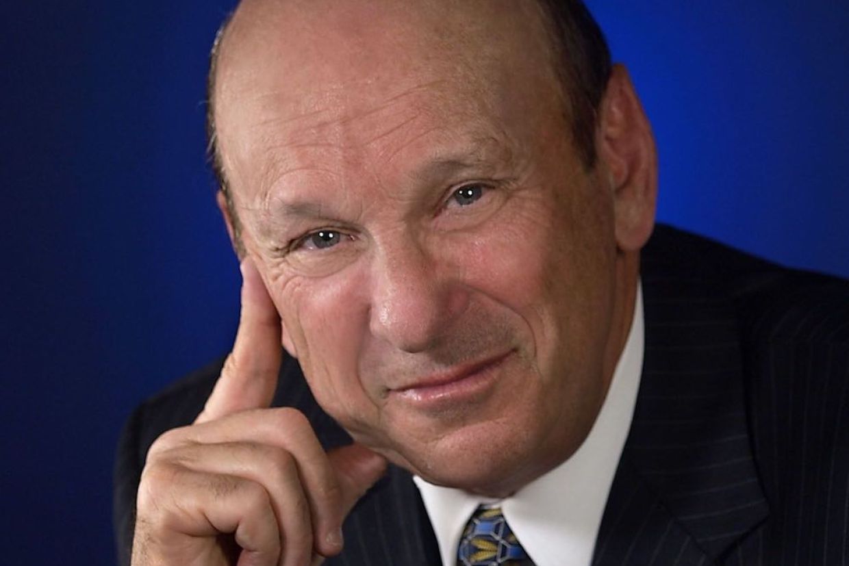 Headshot of Seymour Schulich smiling, wearing a suit and a tie, with a dark blue background.