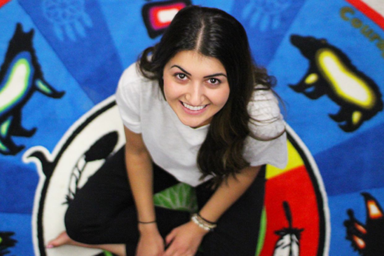 Eleni Fortis, an Indigenous woman, sits cross-legged on an Indigenous designed blanket featuring various animals on a blue background.