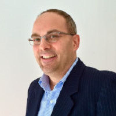 Stacy Kelly smiling he is wearing glasses a light blue shirt and dark blue jacket 