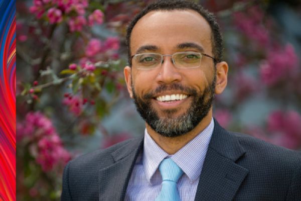 Photo of Edward Thomas looking at the camera smiling. Edward is wearing glasses, a blue shirt, tie and suit jacket