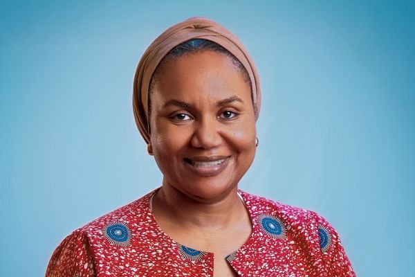 Halima Wali standing against a blue background. They are wearing a red patterned jacket with blue circular designs over a white top and have a beige headscarf.