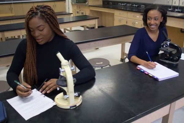 Two women seated at a table in a science lab, focused on their research with various lab tools nearby.