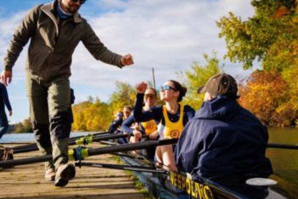 A group of young adults row a boat at the Ontario University Athletics dock, greeting a supporter from the rowing team.