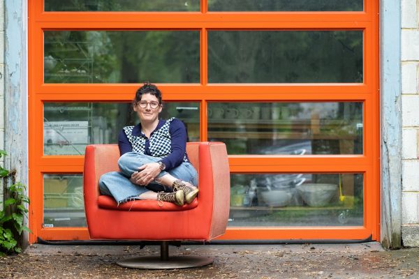 Marney McDiarmid sitting on an orange chair, with legs crossed, outside her studio's orange door.  