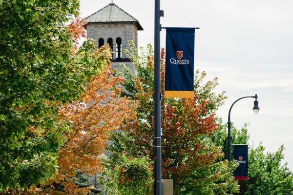 Photo showing the tree line along University Avenue, with Grant Hall in the background a light pole with a Queen's banner in the foreground. 