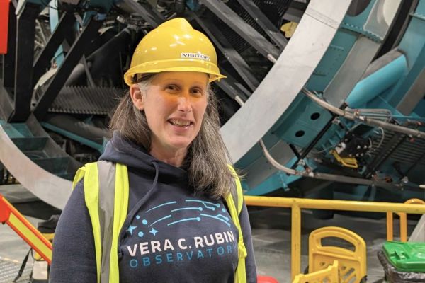 Dr. Melissa Graham, wearing a hard hat and blue shirt, stands before a large machine at the Vera C. Rubin Observatory.