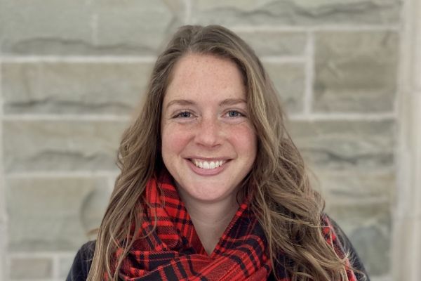 Smiling woman with long brown hair, wearing a red and black plaid scarf, stands in front of a textured stone wall background. Warm and friendly tone.