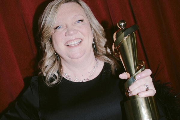 A smiling Jeannie Hunter holding her Juno, with a red curtain behind her. 