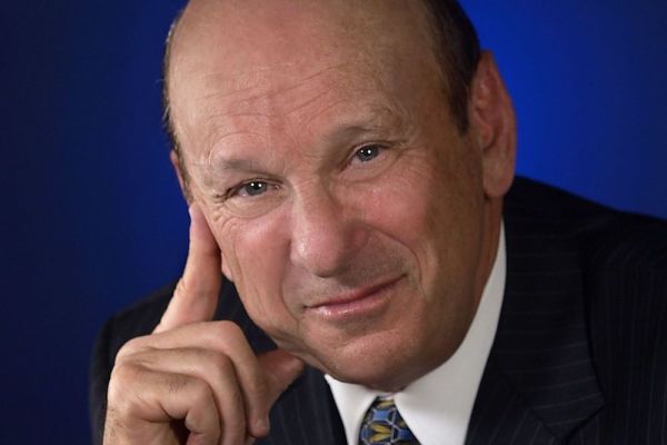 Headshot of Seymour Schulich smiling, wearing a suit and a tie, with a dark blue background.