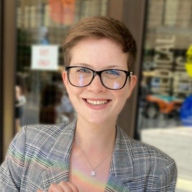 Devon Harvey smiles at the camera wearing a light grey plaid suit and black rimmed glasses.