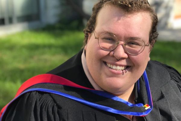 Picture of Wilde, fat person with short cropped brown hair wearing glasses.  They are wearing a Master of Philosophy hood and graduate gown, and there is a green sweater vest underneath the robe. There is green grass and a grey stone building behind them. 