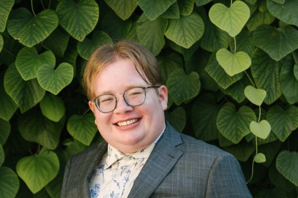 A photograph of Drumlin, a white nonbinary person with short brown hair and glasses. The photo is from the waist up, and they are wearing a grey suitjacket and a white dress shirt with blue flowers on it. The wall behind them is entirely covered in leaves. Photo by Viara Mileva. 
