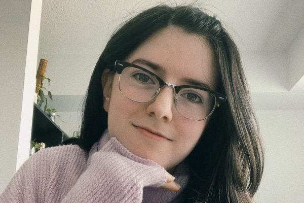 Photo of a young woman with brown hair and glasses. The photo displays her from the shoulders up smiling as her hand rests under her chin.