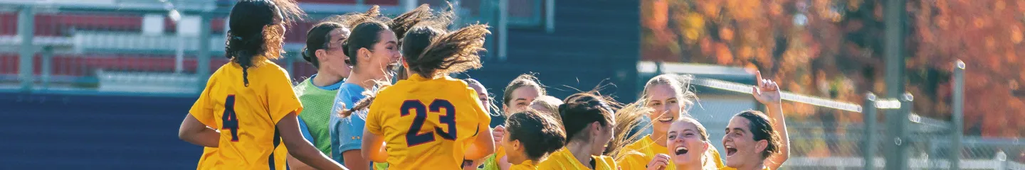 Photo of a group of Queen’s soccer players in yellow uniforms celebrate together in a huddle on an outdoor field.