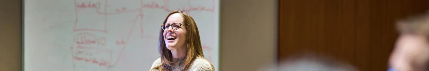 Teacher in front of class with a whiteboard as background.