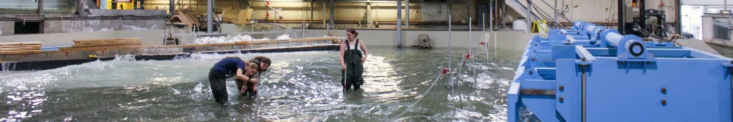 A photo of researchers standing in a large indoor water tank facility while conducting experiments